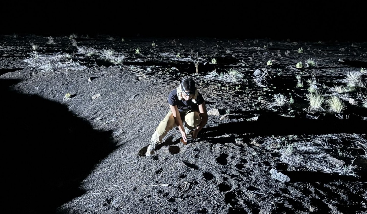 A person wearing boots and protective eye goggles crouches looking at a rock on the ground in low light whilst holding scientific equipment.